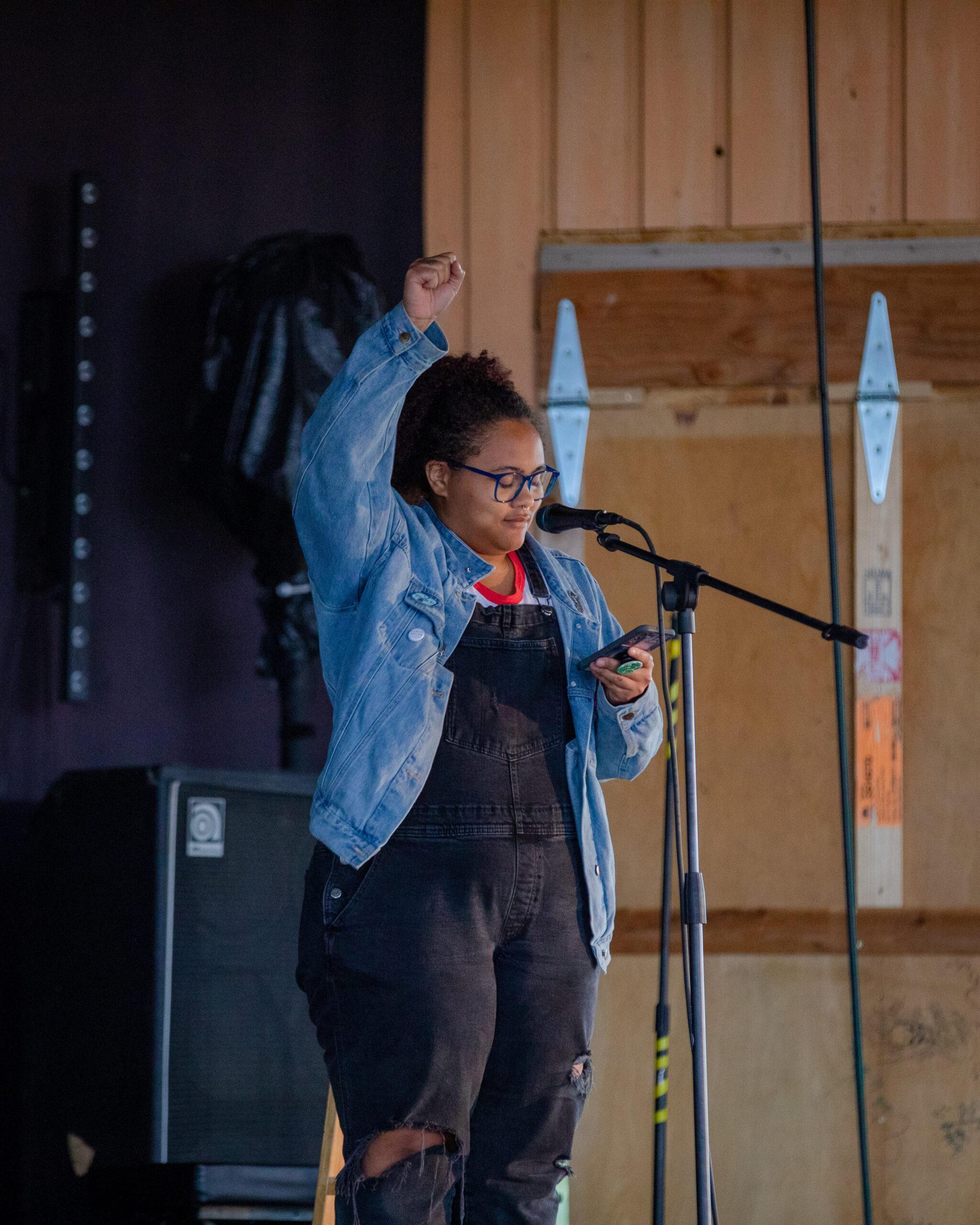 A student reads her creative work on stage while raising her fist at the I Bar Ranch Open Mic Night