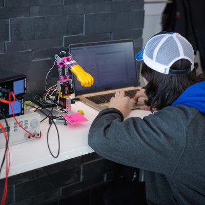 A student codes on his laptop to program a robotic arm next to him.