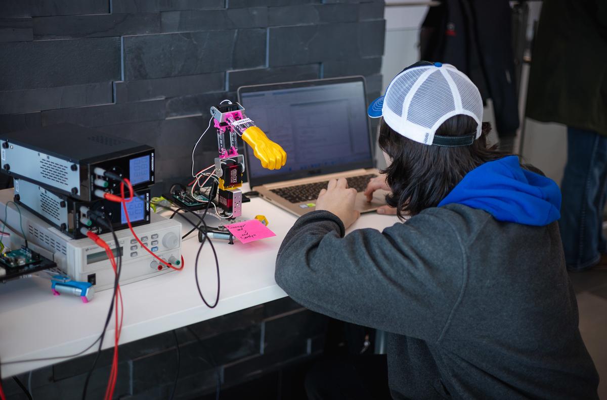 A student codes on his laptop to program a robotic arm next to him.