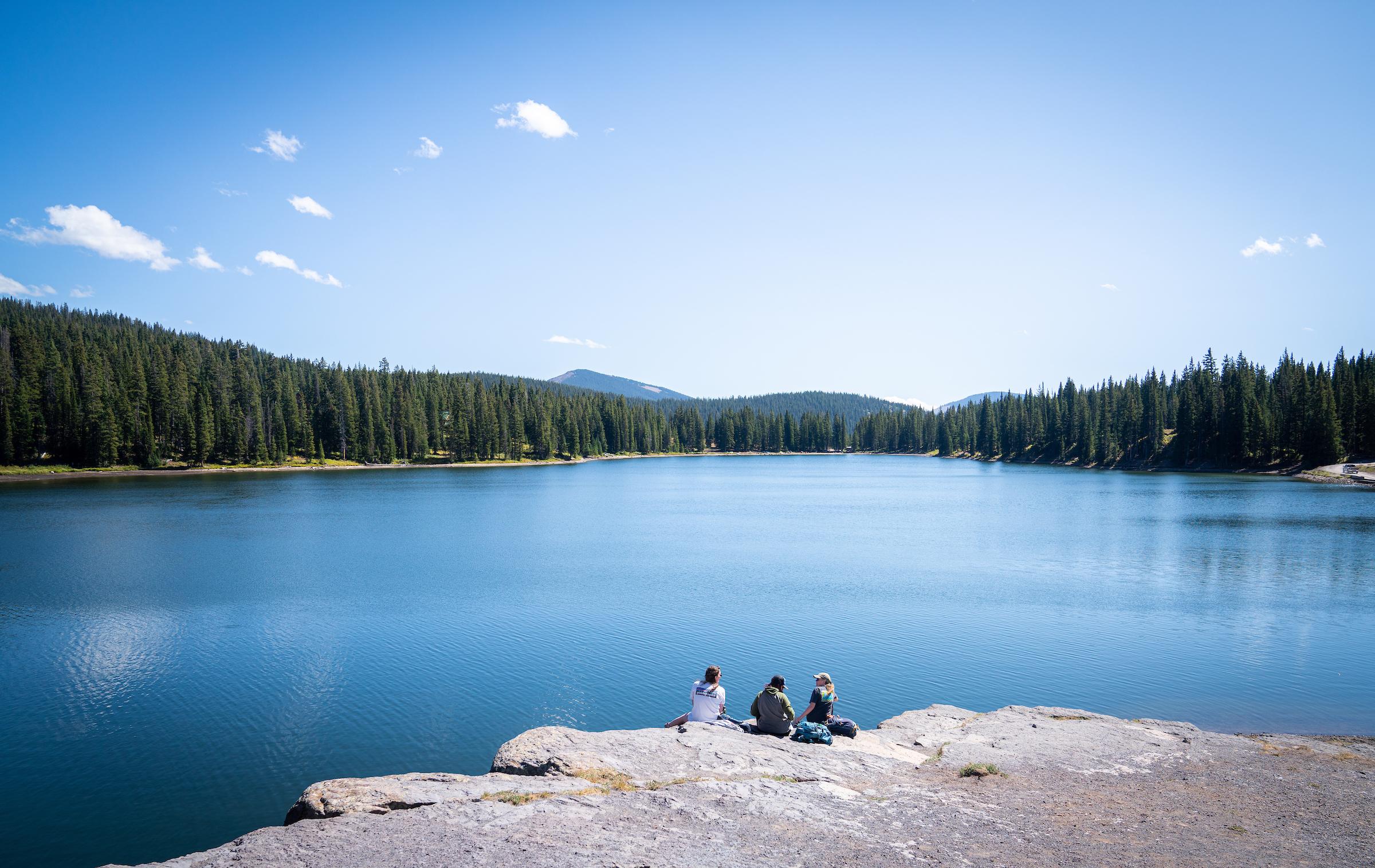 Several students sit near the edge of a mountain lake.