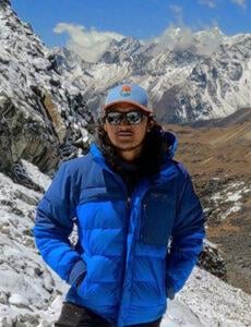 Man wearing a hat, sunglasses, and blue jacket while standing in front of snowy mountains