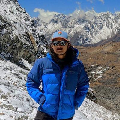 Man wearing a hat, sunglasses, and blue jacket while standing in front of snowy mountains