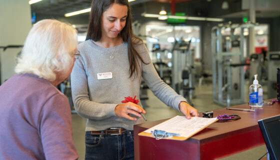 A student explains a fitness program to a community member visiting the gym.