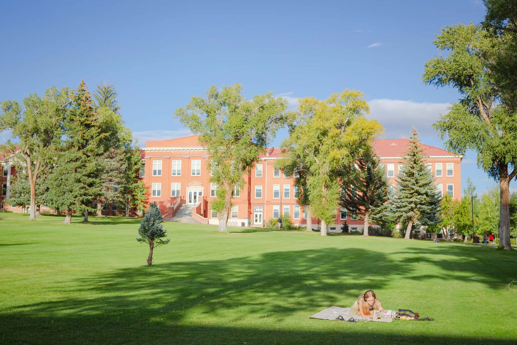 A person relaxes on a blanket in a sunlit grassy area, with a large red brick building and trees in the background under a clear blue sky.