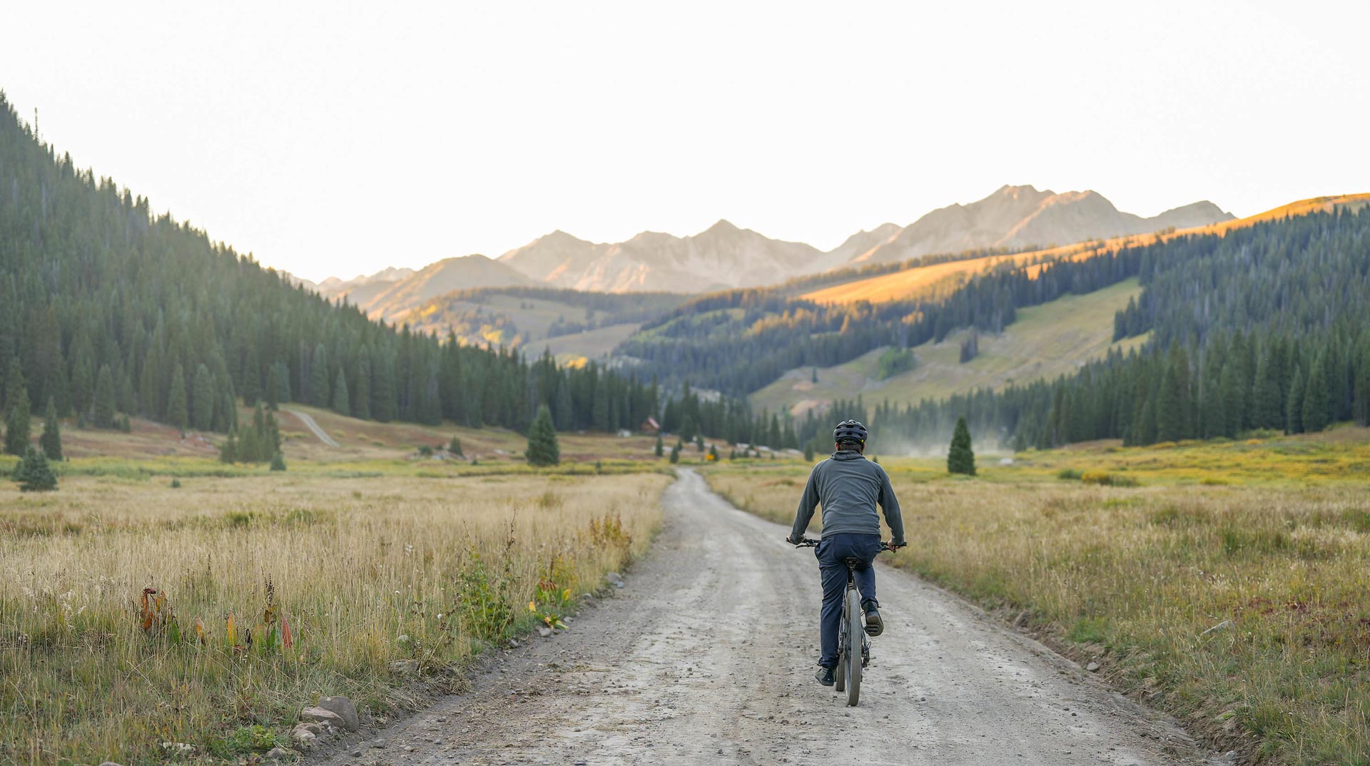 A mountain biker pedals up a dirt road at sunset with mountains and fall colors in the background.