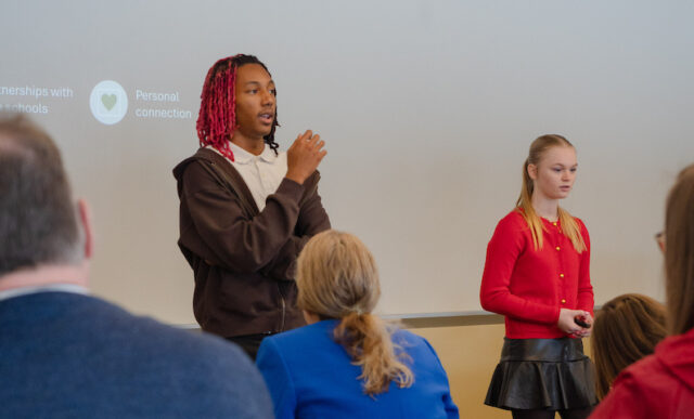 First presenting team, on funding for a college. Young boy with red hair stands adressing the judges with female teammate standing to the right of him .