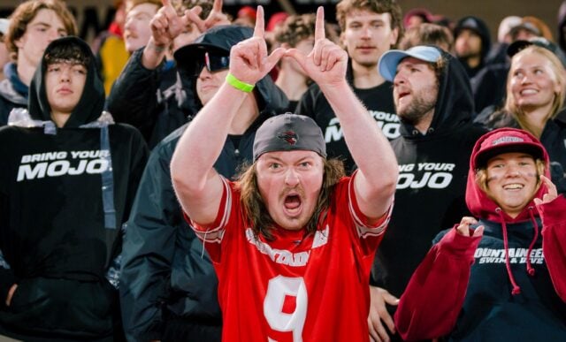 A student in the crow section holds his hands up in a W during a football game.