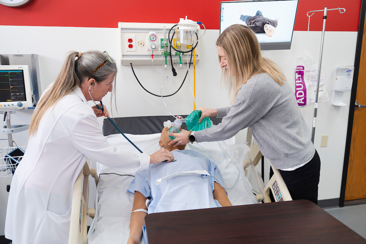 Nursing faculty, Mary Sawyer listens to breathing with a stethoscope while a student practices bagging a mannequin in the simulation lab.