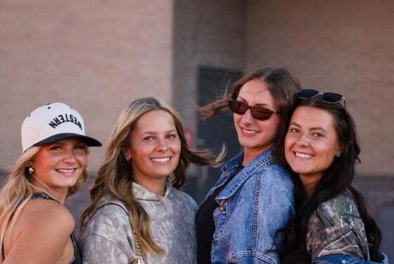 Four friends smile together outdoors, dressed in casual attire including a baseball cap, camouflage, and denim.