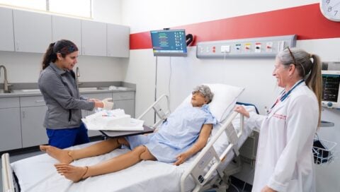 A healthcare professional prepares supplies while another observes as a patient lies on a hospital bed in a simulation room.
