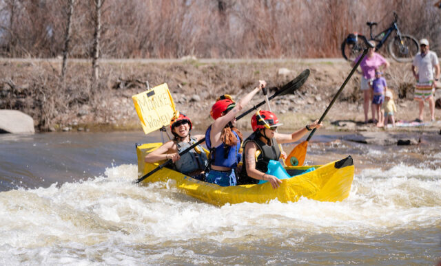 Three female western students in a banana shaped cardboard box foat down the river. They wave their ores in the air cheering to the crowd during the 2025 River Ruckus.