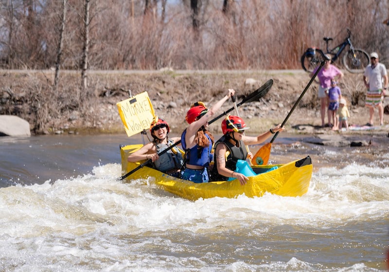 Three female western students in a banana shaped cardboard box foat down the river. They wave their ores in the air cheering to the crowd during the 2025 River Ruckus.