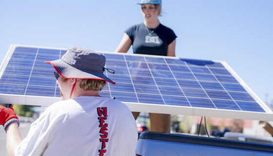 A student stands on a truck bed as she passes solar panels to another student on a blue sky day.