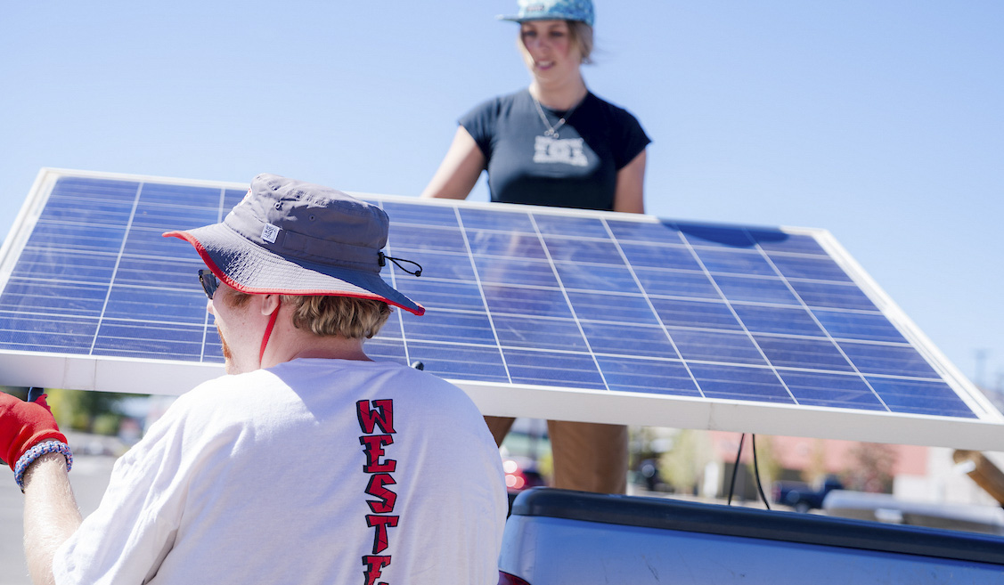 A student stands on a truck bed as she passes solar panels to another student on a blue sky day.