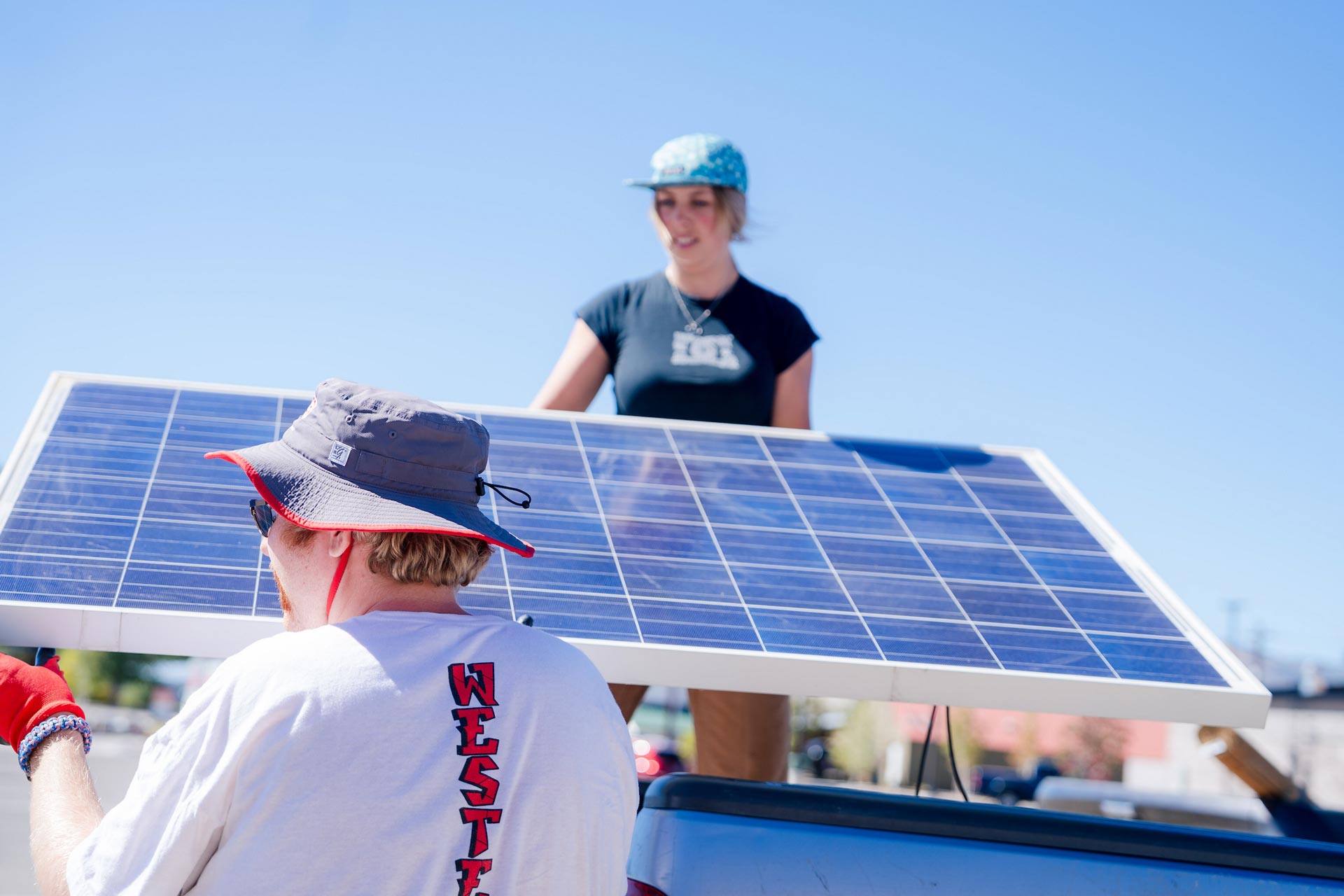 A student stands on a truck bed as she passes solar panels to another student on a blue sky day.