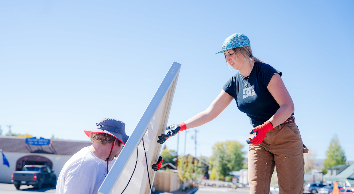 A student stands on a truck bed as she passes solar panels to another student on a blue sky day.