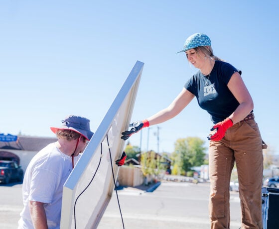 A student stands on a truck bed as she passes solar panels to another student on a blue sky day.
