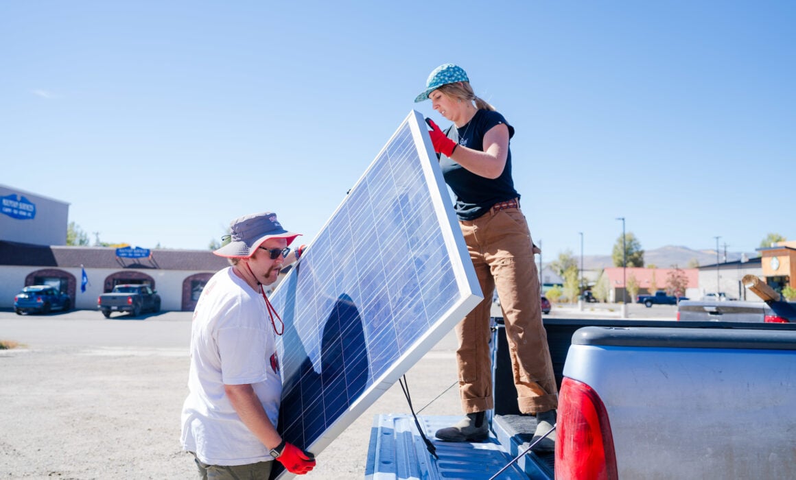 A student stands on a truck bed as she passes solar panels to another student on a blue sky day.