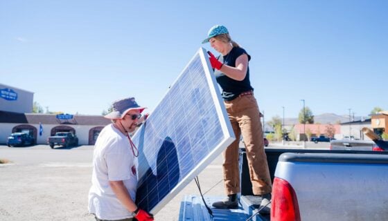 A student stands on a truck bed as she passes solar panels to another student on a blue sky day.