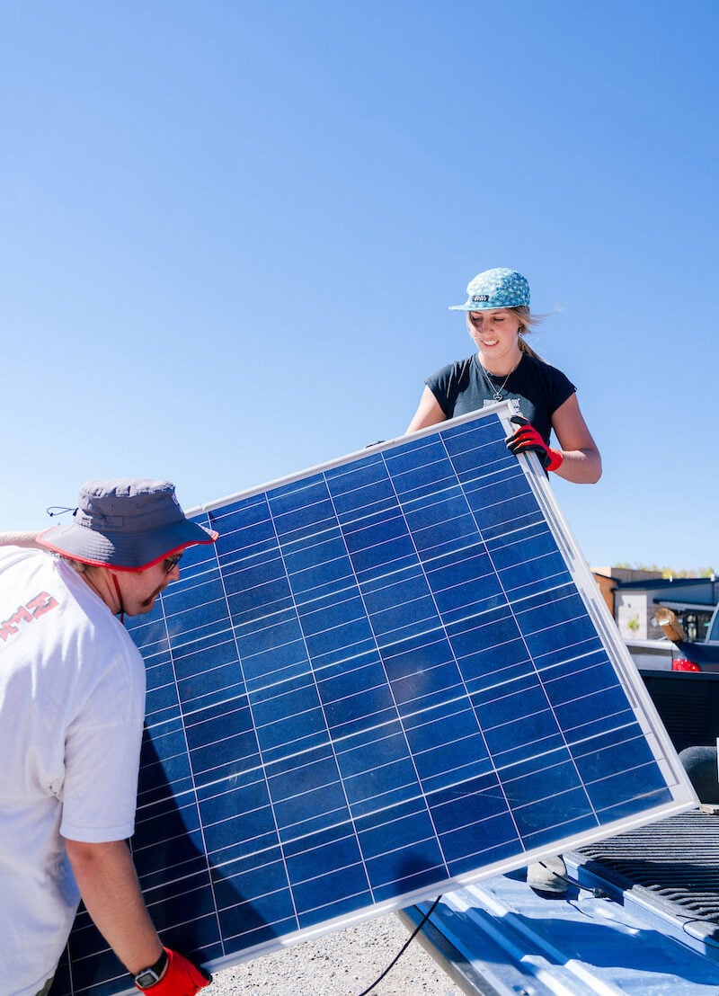 A student stands on a truck bed as she passes solar panels to another student on a blue sky day.