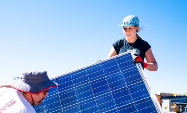 A student stands on a truck bed as she passes solar panels to another student on a blue sky day.
