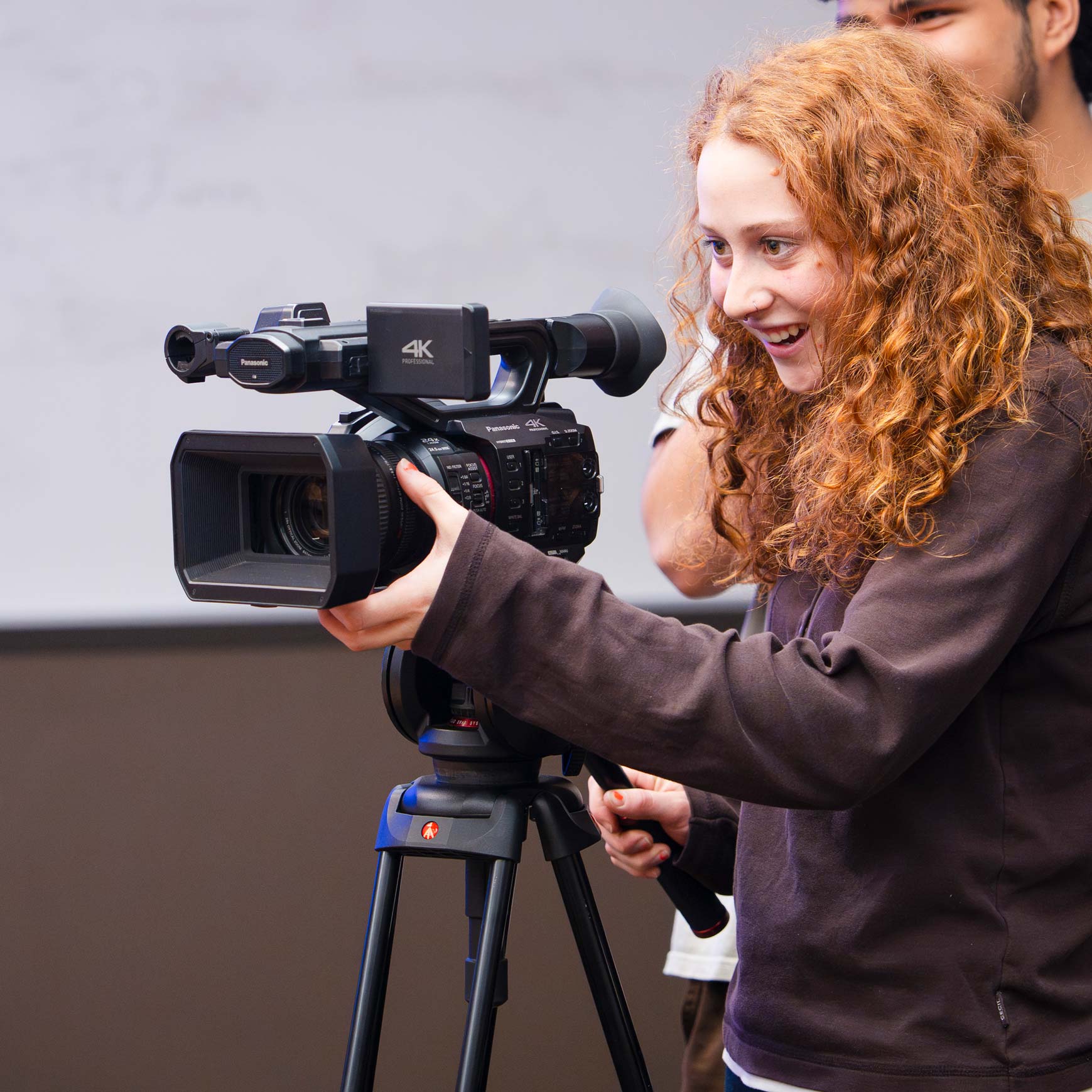 A person with curly hair operates a professional video camera on a tripod while others observe in a classroom setting.