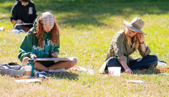 A student sits on grass and watercolor paints a scene on her lap with professor Tina Butterfield sitting next to her.