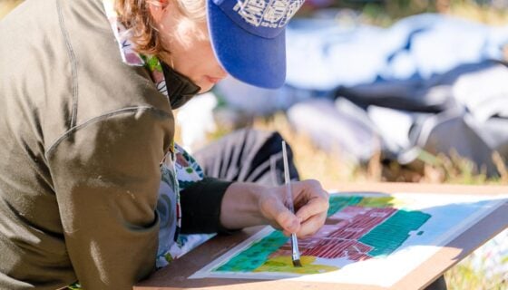 A close up of a student sittin gon grass and painting a barn with watercolors.