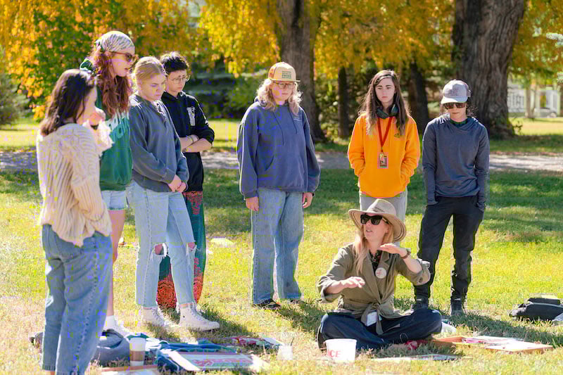 A small art class stands on grass around professor Tina Butterfield as she explains a watercolor painting technique.