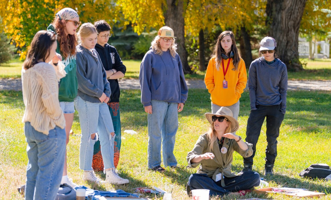 A small art class stands on grass around professor Tina Butterfield as she explains a watercolor painting technique.