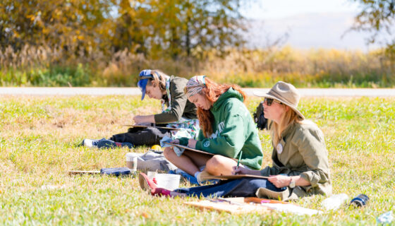 Two students and a professor sit on the grass and watercolor paint a scene with their canvases resting on their laps.