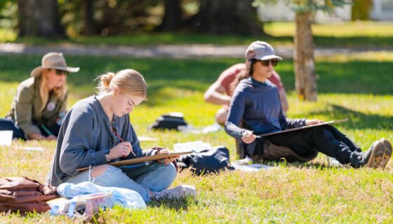 Two students sit on grass and watercolor paint a scene with their canvases on their laps.