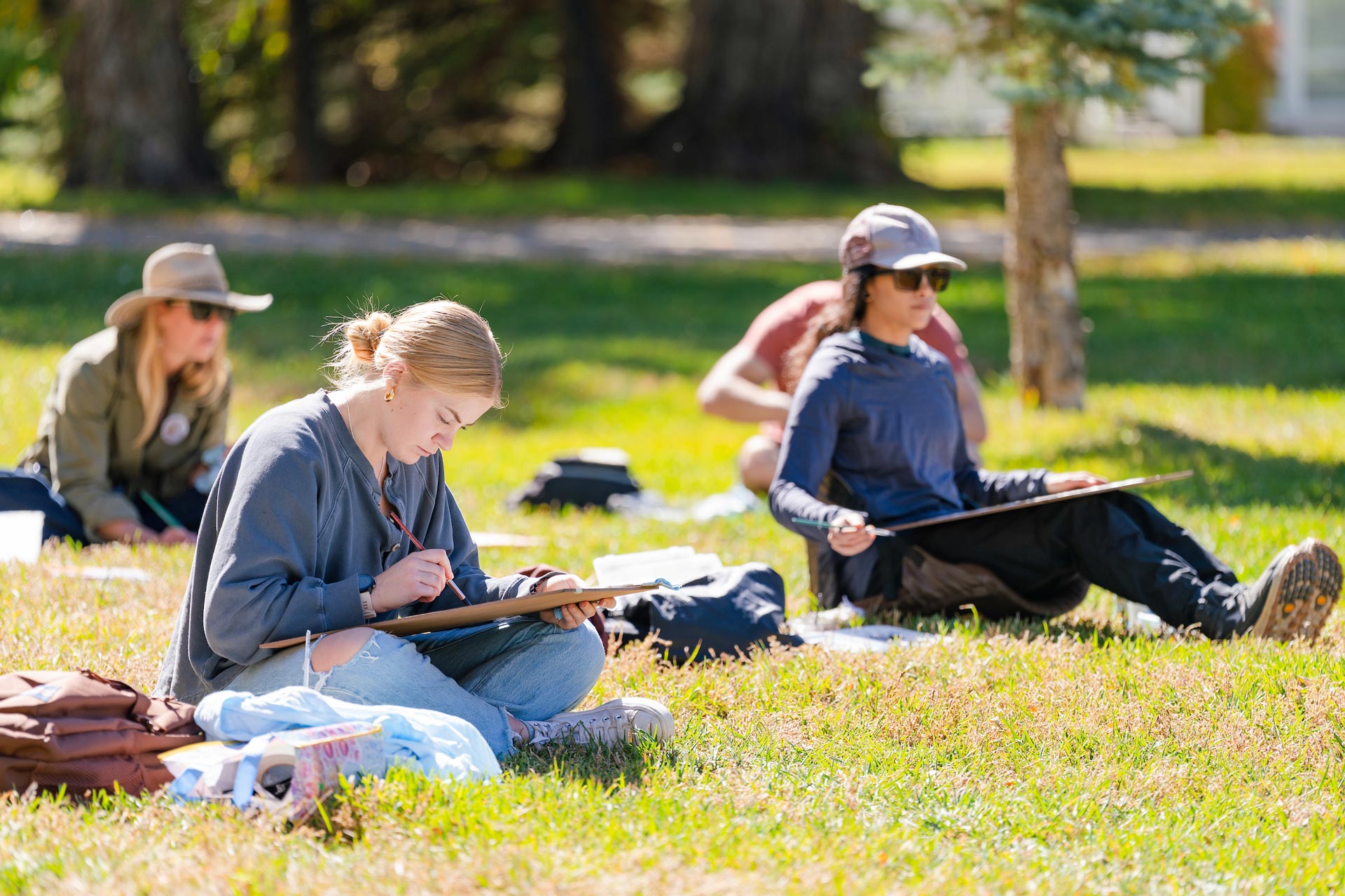 Two students sit on grass and watercolor paint a scene with their canvases on their laps.