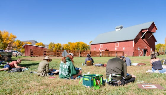A small art class sits on the grass and watercolor paints a barn that is in front of them.