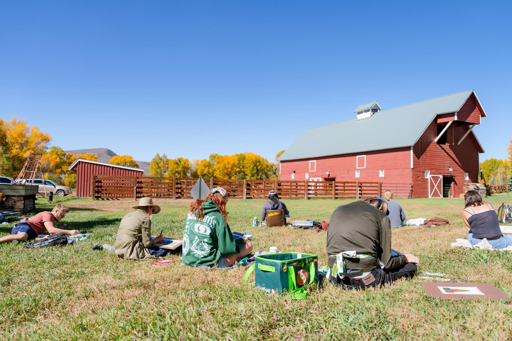 A small art class sits on the grass and watercolor paints a barn that is in front of them.
