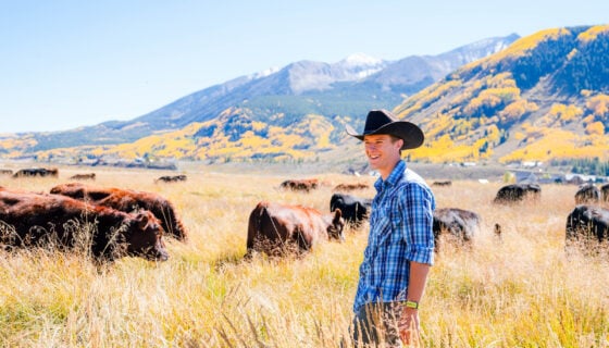 A graduate student wearing a cowboy hat smiles as he stands on Verzuh Ranch wetlands with cattle and Whetstone Mountain behind him.