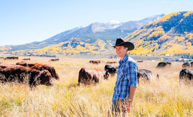 A graduate student wearing a cowboy hat smiles as he stands on Verzuh Ranch wetlands with cattle and Whetstone Mountain behind him.