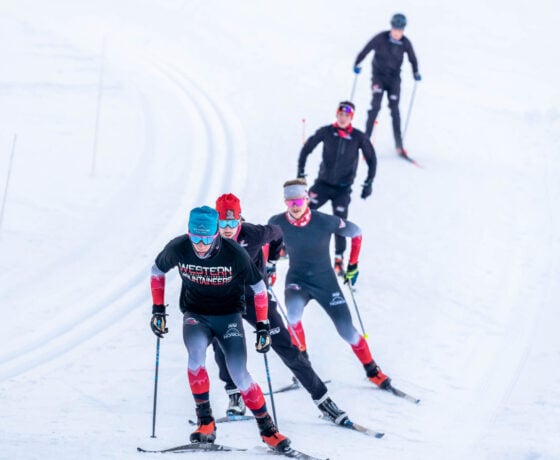 Five Nordic skiers wearing their uniforms skate in a single file line during practice.