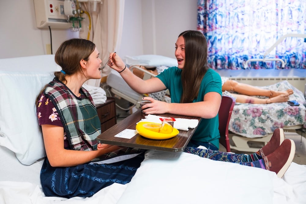A student feeds another student (who is seated on a bed) a spoonful of chocolate pudding while she practices the procedure for feeding a patient during a CNA course.