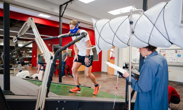 A student wears a face mask as he walks on an inclined treadmill inside the HAP Lab, simulating a trek to Mt. Everest base camp. A different student holds a clipboard and collects data.