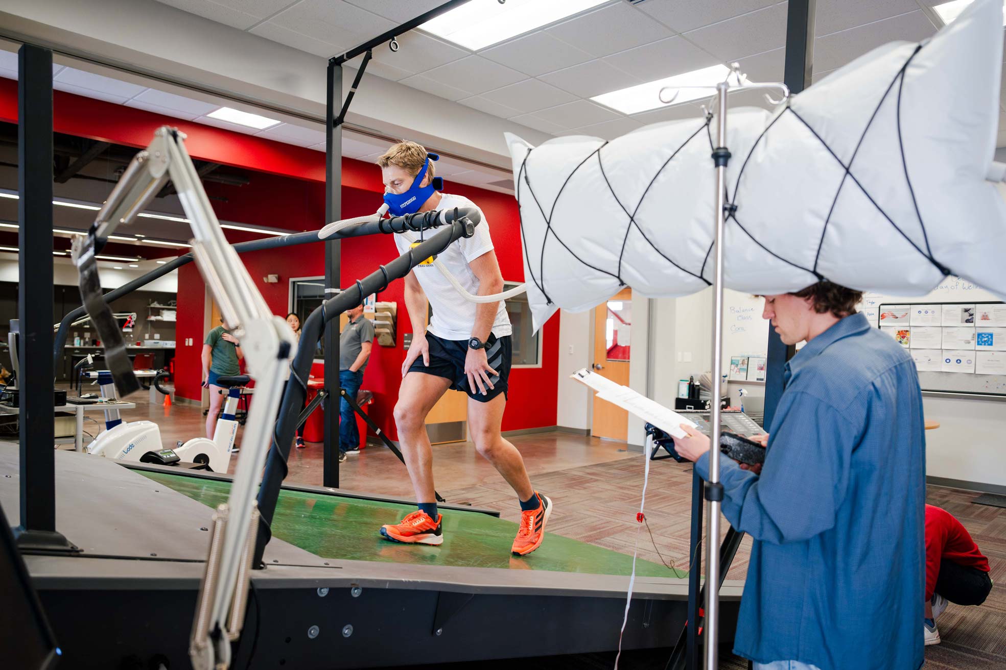 A student wears a face mask as he walks on an inclined treadmill inside the HAP Lab, simulating a trek to Mt. Everest base camp. A different student holds a clipboard and collects data.