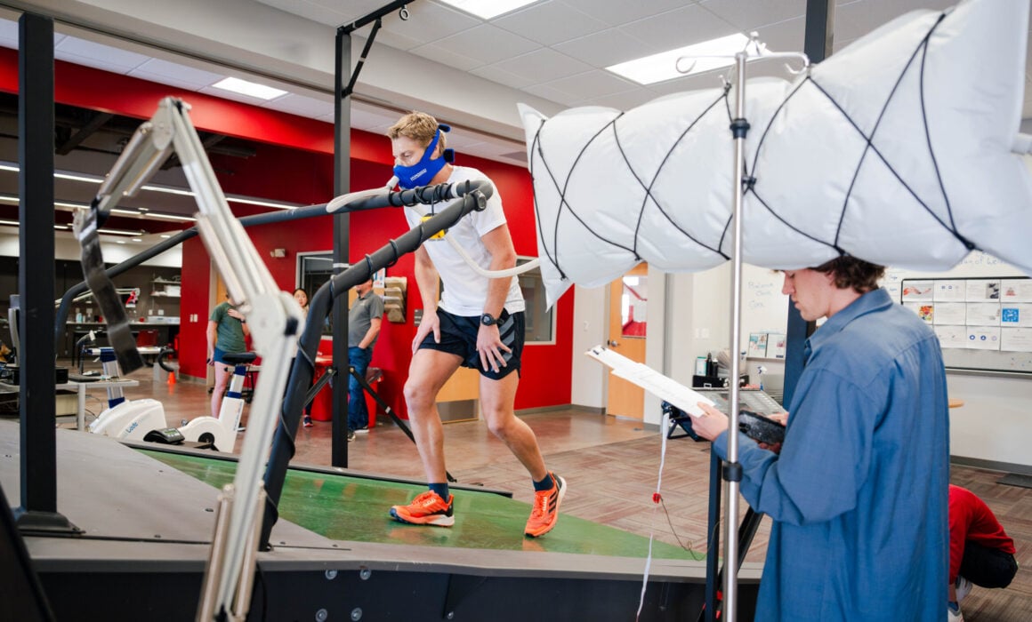 A student wears a face mask as he walks on an inclined treadmill inside the HAP Lab, simulating a trek to Mt. Everest base camp. A different student holds a clipboard and collects data.