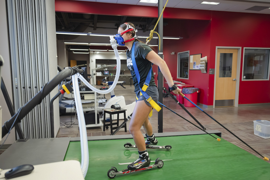 A Mountain Sports Nordic Skiing athlete roller skis on a treadmill in the HAP Lab. She is wearing a face mask that is measuring her VO2 Max.