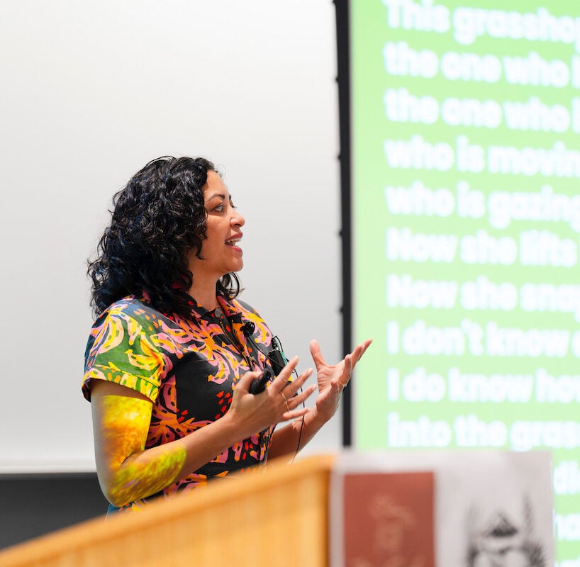 A person with curly hair speaks at a podium in front of a projection screen displaying text.