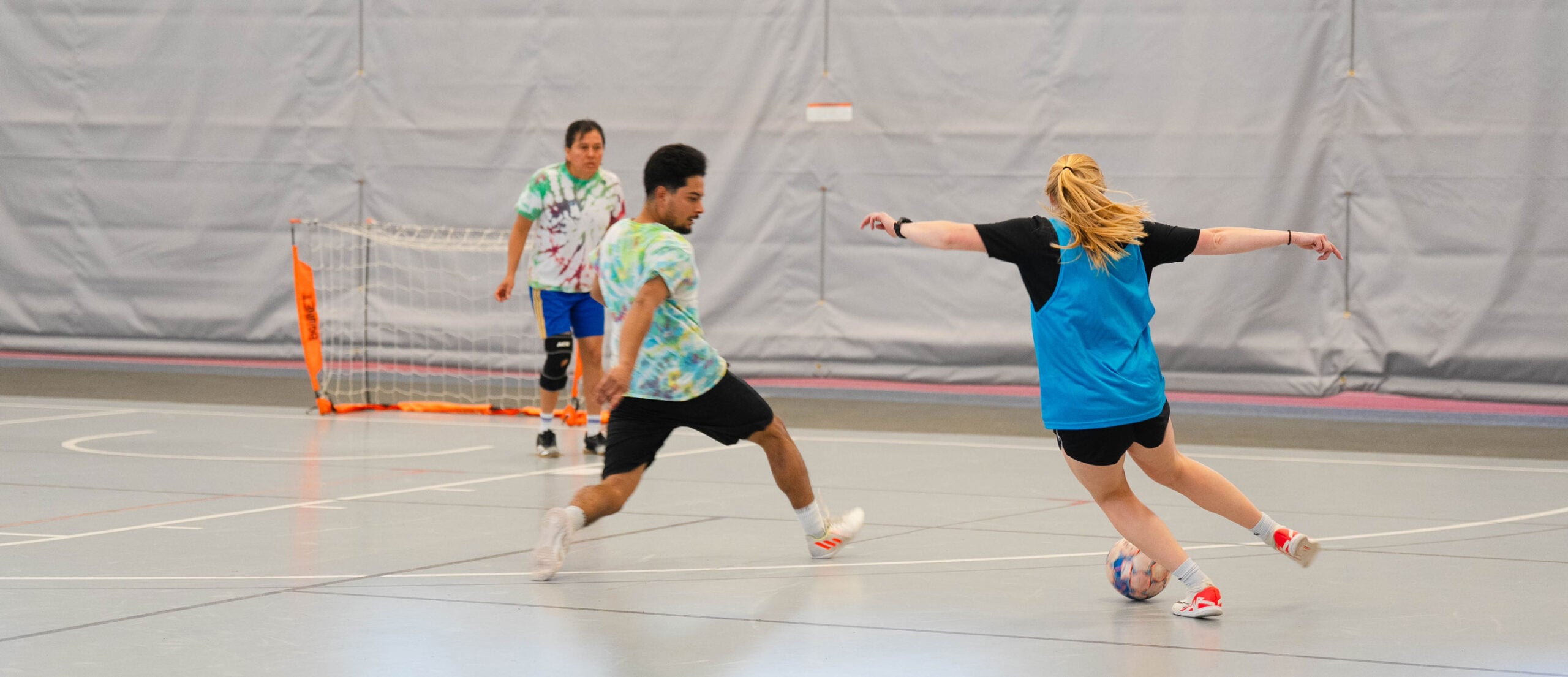 Three players are actively engaged in an indoor soccer game, with one player controlling the ball while another defends and a third watches from the sidelines.
