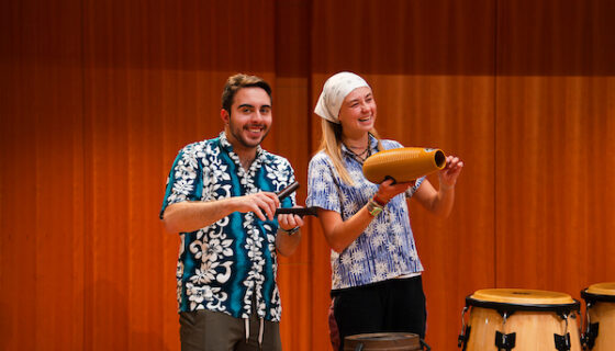 Two students smile while playing some hand held percussion instruments during a steel band practice in the Kincaid Concert Hall.
