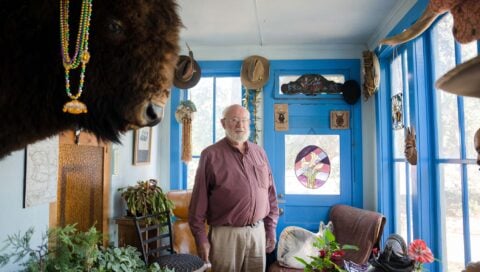 A photo of Butch Clark standing in one of the rooms of his house that has many windows and a large bison head mounted on the wall.
