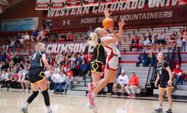 A basketball player in a white and red uniform jumps to shoot the ball while being defended by an opponent in a black and yellow uniform, with another player observing from the sideline during a game at Western Colorado University.