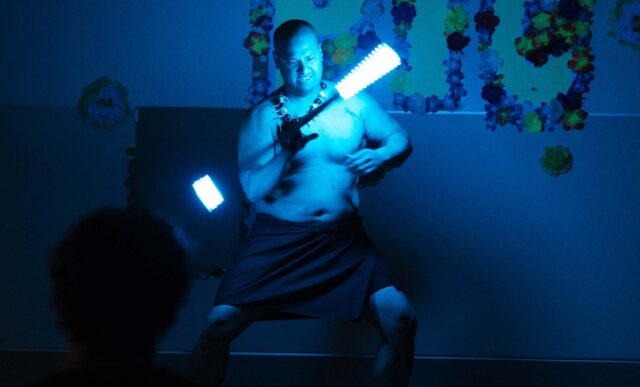 A performer on stage under blue lighting holds glowing sticks, with a banner reading "Asian & Pacific Islanders Club: The Līlāu!" behind them.