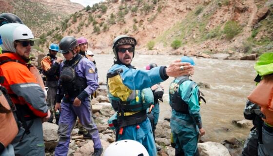 A group of river rescuers in safety gear stand on rocky riverbank, with one person gesturing toward the water as they prepare for a rescue drill.